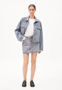 Veste en denim bleu clair à coupe oversize, associée à une jupe en denim bleu clair. Le modèle porte un t-shirt blanc et des mocassins noirs.