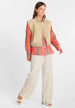 Donna che indossa un gilet beige senza maniche sopra una camicia vivace con stampa tigre, pantaloni ampi a righe crema e tacchi bianchi aperti in punta, in piedi di profilo.