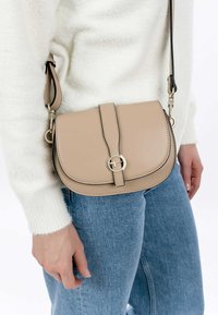 Borsa a tracolla in pelle beige dalla forma arrotondata, con hardware dorato, chiusura a patta e tracolla nera regolabile, tenuta contro un maglione bianco.