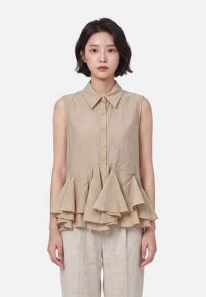 Donna con top beige senza maniche con bottoni e orlo peplo arricciato e pantaloni larghi chiari, in piedi su sfondo bianco.