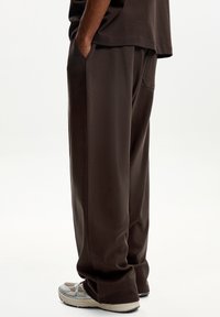 Rjavi bombažni hlače z ohlapnim krojem, teksturirano tkanino, stranskimi žepi in ravnim krojem nogavic, v kombinaciji s sivimi športnimi čevlji.