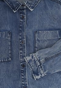 Lichtblauwe denimblouse met een puntige kraag, twee borstzakken, zwarte knopen en een licht verweerde textuur.