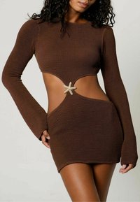 Femme portant une robe mini à manches longues en tricot texturé marron avec une taille découpée et un fermoir en forme d'étoile de mer au centre.