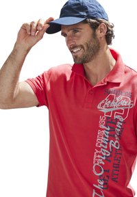 Rotes Poloshirt mit Kragen, das auf der linken Seite mit einem grafischen Design in Weiß und Blau versehen ist; getragen mit einer marineblauen Kappe.