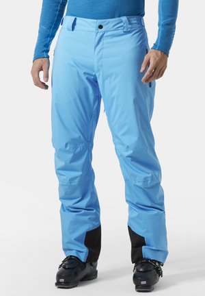 Mann trägt hellblaue Skihose mit schwarzen verstärkten Bündchen über schwarzen Skischuhen, steht mit den Händen an den Seiten vor einem schlichten Hintergrund.