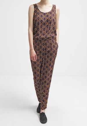 Person iført ærmeløs jumpsuit med geometrisk diamantmønster i mørke farver og sorte sneakers, stående med den ene hånd i lommen.