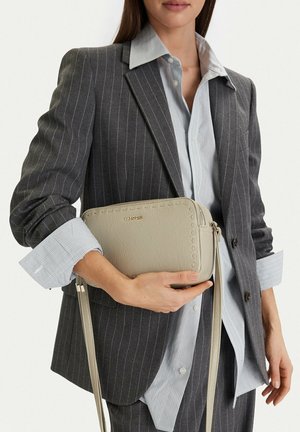 Mujer con blazer gris a rayas y camisa sosteniendo un bolso cruzado beige con el logo visible de la marca Lasocki.