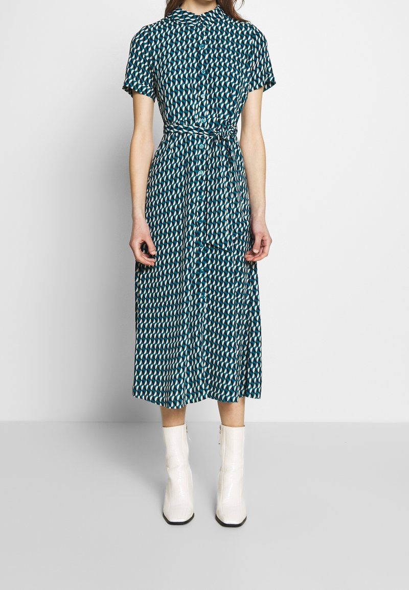 Femme portant une robe midi boutonnée à motif turquoise, à manches courtes, et des bottines blanches, debout devant un fond blanc uni.