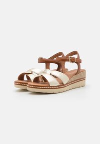 Sandalias cuña con parte superior metálica dorada y acentos de cuero marrón. Cuenta con una correa ajustable y un nudo decorativo en la parte delantera.