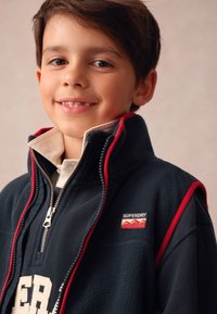 Tamsiai mėlynas flisinis striukė su užtrauktuku, raudonais akcentais, aukštu apykakliu ir siuvinėtu „Superdry“ logotipu ant krūtinės. Po ja baltas sluoksniuotas marškinėlis.