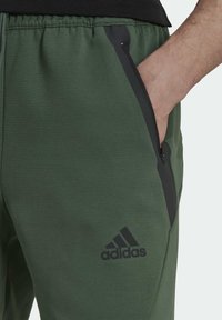 Zelené joggingové kalhoty vyrobené z měkkého materiálu, s černými zipovými kapsami a logem Adidas. Design zahrnuje elastický pas.