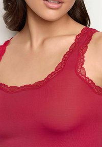 Rød tanktop lavet af blødt stof med en blondekant langs udskæringen og stropperne. Designet er tætsiddende og glat, hvilket fremhæver brystområdet.