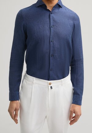 Homme portant une chemise bleu marine à manches longues boutonnée et rentrée dans un pantalon blanc plissé taille haute avec un bouton en bois.