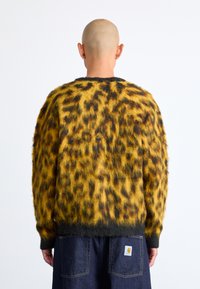 Κίτρινο και μαύρο πουλόβερ με λεοπάρ σχέδιο, φτιαγμένο από μαλακό υλικό, με σκούρα ζώνη στο κάτω μέρος και μακριά μανίκια; φορεμένο πάνω από σκούρα τζιν.