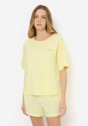 Frau trägt ein locker sitzendes hellgelbes T-Shirt und passende gelb-weiße gestreifte Shorts, steht vor weißem Hintergrund.