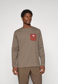 Camisa de manga larga en tejido marrón, con un bolsillo en el pecho color burdeos que tiene una pequeña etiqueta. El diseño incluye un escote redondo y mangas ajustadas.