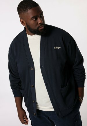 Cardigan blu navy con chiusura a bottoni, due tasche e logo ricamato. Indossato su una camicia bianca, con maniche lunghe e polsini a costine.