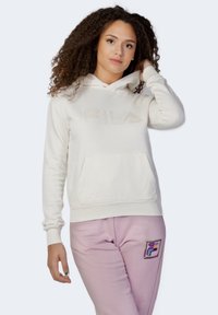 Felpa color crema con cappuccio, dotata di tasca frontale a marsupio e logo "FILA" in rilievo. Polsini e vita a costine. Abbinata a pantaloni della tuta rosa.