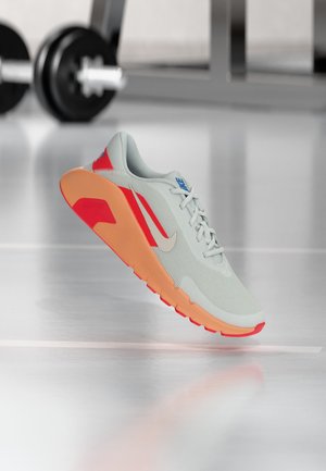 Hellgrauer und oranger Nike-Laufschuh mit roten Akzenten, der auf einem reflektierenden Fitnessstudio-Boden schwebt, mit einer verschwommenen Hantel im Hintergrund.