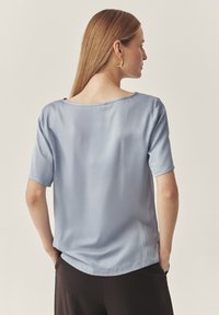 Hellblaues Kurzarmoberteil aus glattem Stoff, mit weit ausgeschnittenem Hals und lockerer Passform, abgerundetem Saum und subtiler Schattierung.
