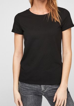 Femme portant un t-shirt noir uni à manches courtes et un jean gris foncé, debout devant un fond clair uni.
