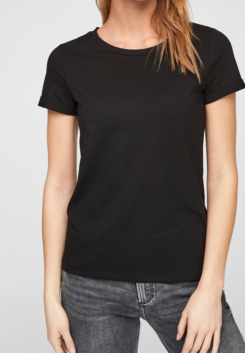 Femme portant un t-shirt noir uni à manches courtes et un jean gris foncé, debout devant un fond clair uni.