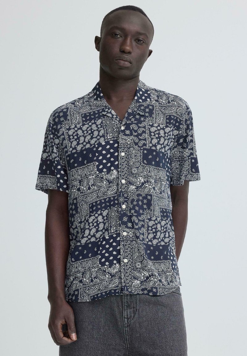 Homme portant une chemise bleu marine à manches courtes avec des motifs cachemire blancs et des motifs patchwork, debout devant un fond gris clair.