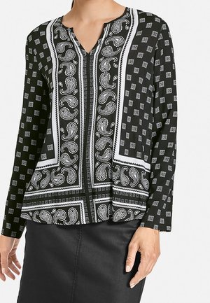 Femme portant un chemisier noir à manches longues avec des motifs cachemire blancs et géométriques, assorti à une jupe noire.