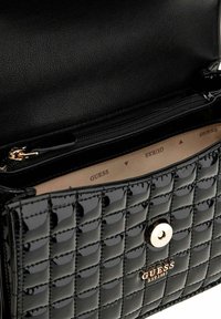 Sac à main en cuir verni matelassé noir avec quincaillerie dorée, doté d'un fermoir à pression et du logo "GUESS" embossé à l'avant.