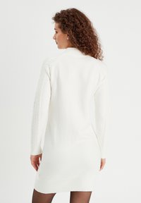 Robe pull blanche en tricot avec des détails en côtes sur les manches, silhouette ajustée et texture douce. La longueur atteint le milieu de la cuisse, encolure ras du cou.