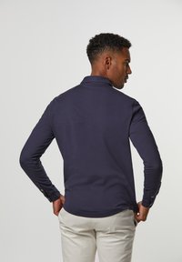 Mann in einem dunkelblauen Langarmshirt und hellbeigen Hosen mit Händen in den Hüften, abgewandt vor einem einfarbigen hellgrauen Hintergrund.