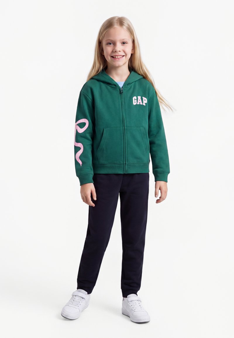 Jeune fille souriante, portant un sweat à capuche zippé vert avec le logo "GAP", un pantalon noir et des baskets blanches, debout sur un fond blanc uni.