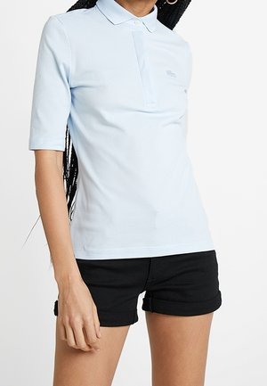 Vrouw draagt een lichtblauw poloshirt met korte mouwen en zwarte opgerolde shorts tegen een effen witte achtergrond.