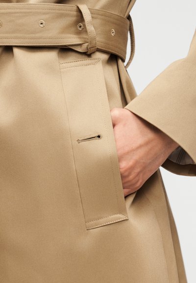 Χέρι που έχει τοποθετηθεί στην πλαϊνή τσέπη ενός μπεζ καμπαρντίνας με ζώνη στο ίδιο χρώμα και λεπτομέρειες με κουμπότρυπες.