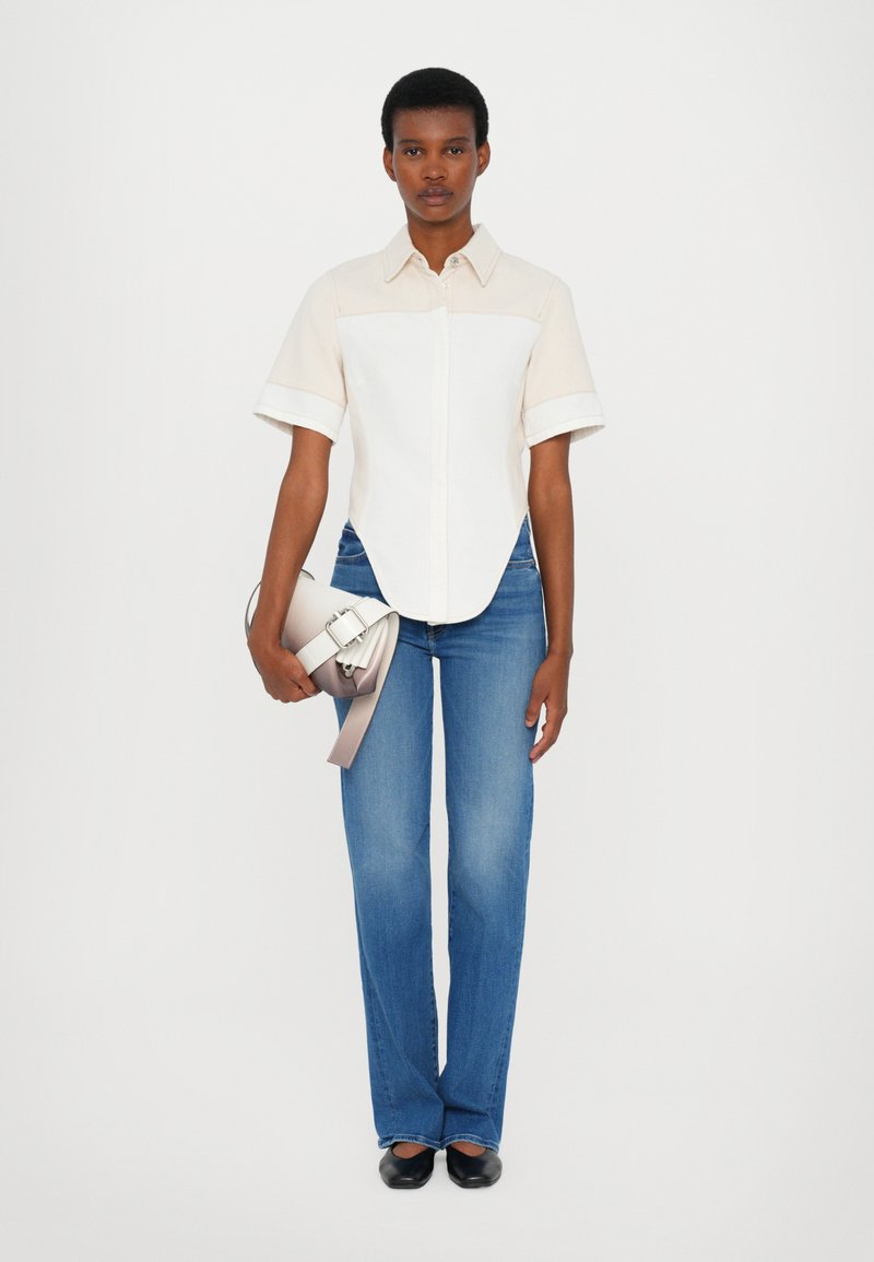 White and beige short-sleeve shirt, blue flared jeans, black flat shoes, holding a gray handbag with a ribbon detail. Simple design.