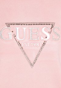 Rosa Stoffhemd mit dem Schriftzug "GUESS" in Silber und Strassverzierungen sowie dem Text "EST. 1981" darunter in weißem Garn.