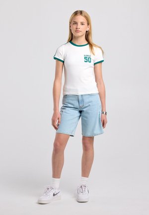 Jeune personne debout, portant un t-shirt blanc avec une bordure verte et le texte "Hawaii 50", un short en jean clair, des chaussettes blanches et des baskets blanches Nike.