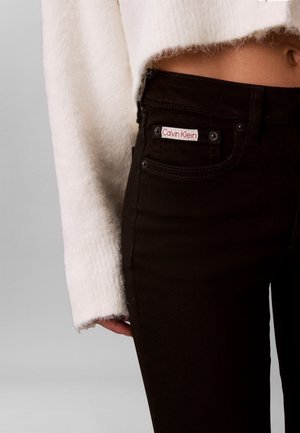 Personne portant un jean noir Calvin Klein et un pull court blanc duveteux, montrant la taille et la main sur un fond gris.