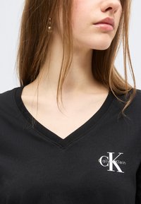 Νεαρή γυναίκα με ίσια ανοιχτόκαφέ μαλλιά που φοράει μαύρο μπλουζάκι Calvin Klein με λευκό λογότυπο στο στήθος.