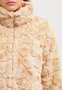 Faux fur-jacka i beige med en texturerad, plysch yta som har ett unikt mönster. Hög krage, mjuk att ta på.