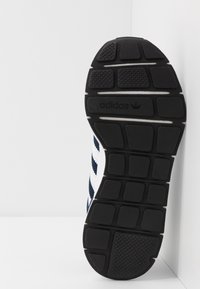Schwarze Gummisohle des Adidas Sneakers mit strukturiertem Profil und Adidas-Logo, vor einem weißen Wandeck-Hintergrund.
