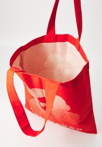 Sac fourre-tout en tissu rouge avec deux poignées robustes, présentant un design floral clair à l'intérieur. Construction simple et fonctionnelle.