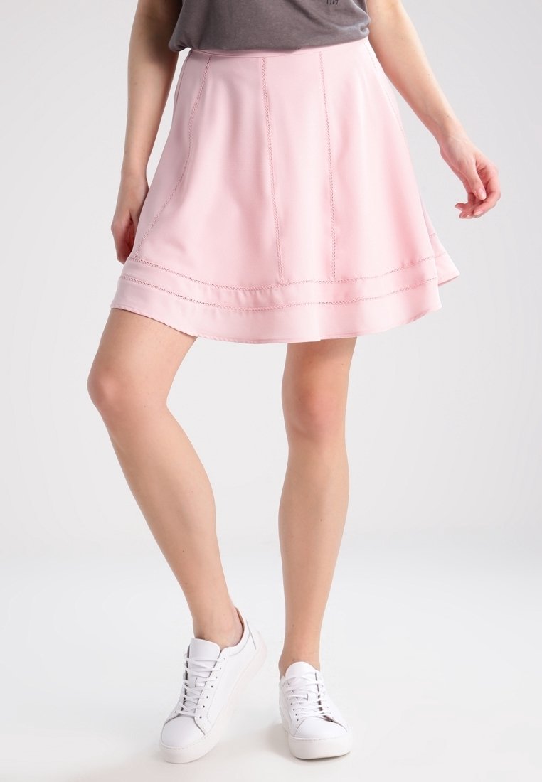 Femme portant une jupe trapèze rose clair avec des détails cousus et des baskets blanches, debout, jambes croisées, sur un fond uni.