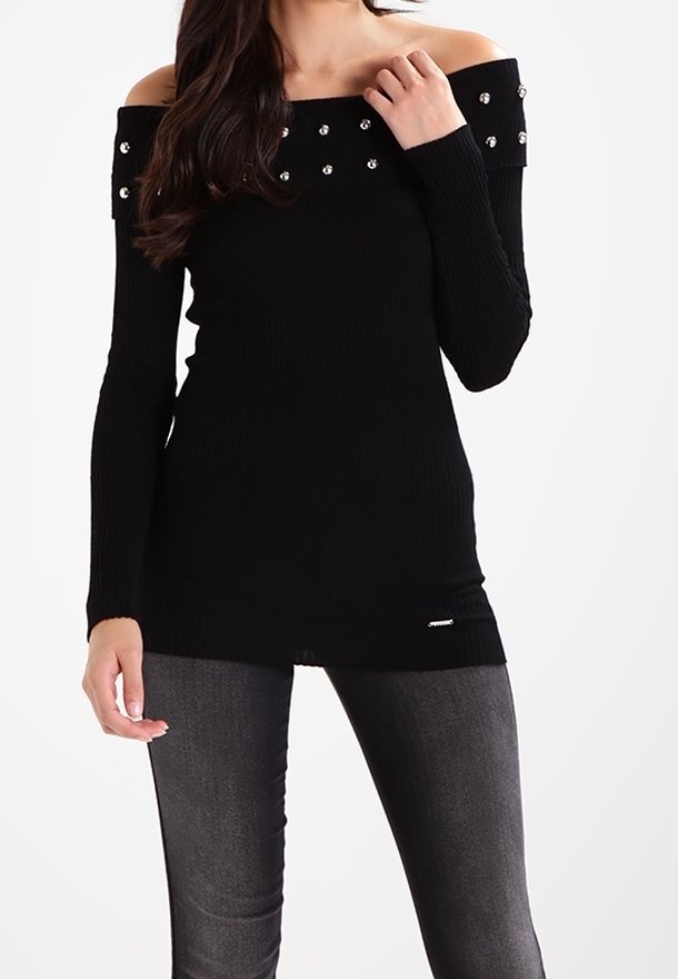 Femme portant un pull noir à épaules dénudées orné de perles, associé à un pantalon ajusté gris foncé.