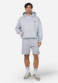 Sweatshirt cinza com capuz e bolso frontal, combinado com calções cinzentos. Apresenta um logo em ambos os itens. Usado com sapatilhas brancas e cinzas.