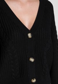 Svart stickad cardigan med V-ringning, texturerat kabelmönster och knappar i guldton nedtill. Slätt, tjockt tyg.