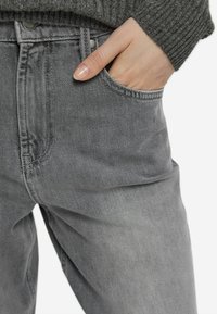 Grå denimjeans med en avslappnad passform, med synliga sömmar, en metalldetaljerad knapp och en framficka med en delvis inskjuten hand.