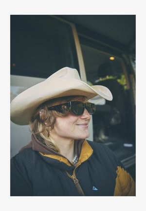 Hellfarbener Cowboyhut, schwarze eckige Sonnenbrille und eine dunkle Jacke mit gelben Akzenten. Lockiges Haar texturiert, geschichtet mit einer braunen Kapuzenjacke.