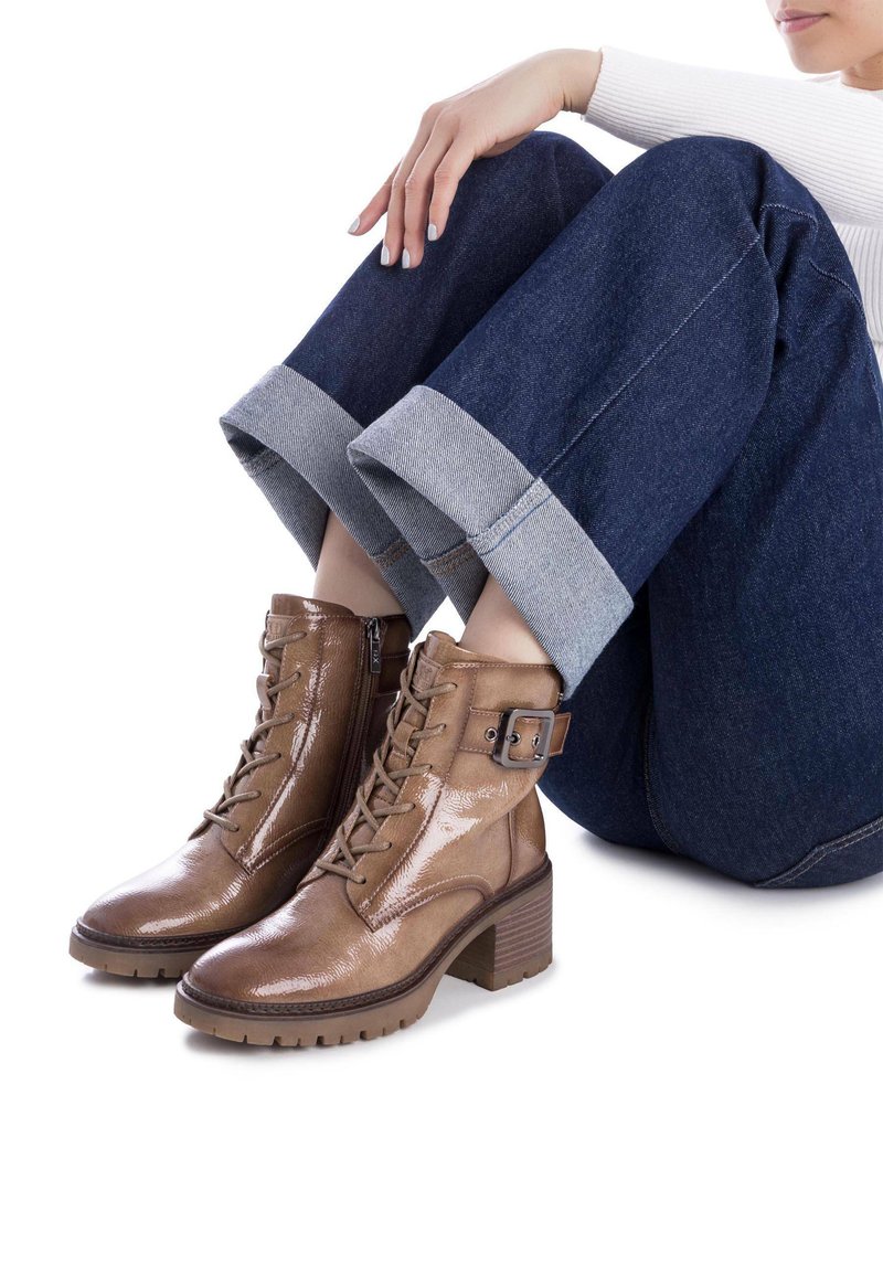 Botas de tobillo de cuero marrón con un acabado brillante, diseño de cordones, cremallera lateral y acento de hebilla metálica. Tacón grueso resistente y suela texturizada.