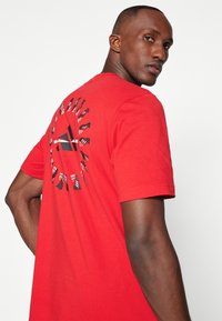 Camiseta de algodón roja con un estampado gráfico en la parte posterior que presenta elementos en blanco y negro y rayas. Diseño de mangas cortas y cuello redondo.
